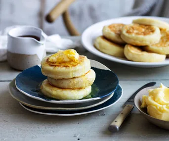 Crumpets on a plate topped with butter and honey, additional crumpets, butter in a dish, and a jug in background.