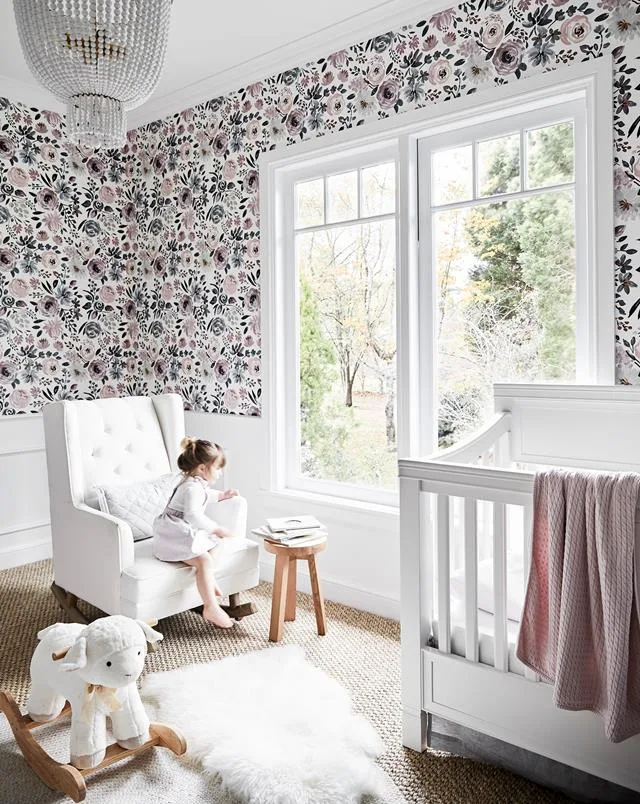 A children's nursery with floral patterned wallpaper, a large window and white nursery furniture. 