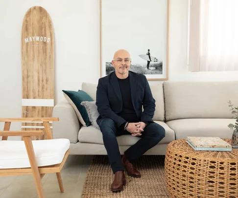 Man sitting on a sofa in a modern living room with a surfboard, framed photo, and a wicker coffee table.