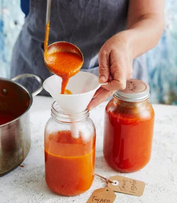 A person is pouring homemade tomato sauce from a ladle through a funnel into a glass jar, with another filled jar nearby.