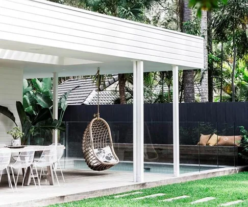 Covered patio with white furniture, hanging wicker chair, and greenery in the background.