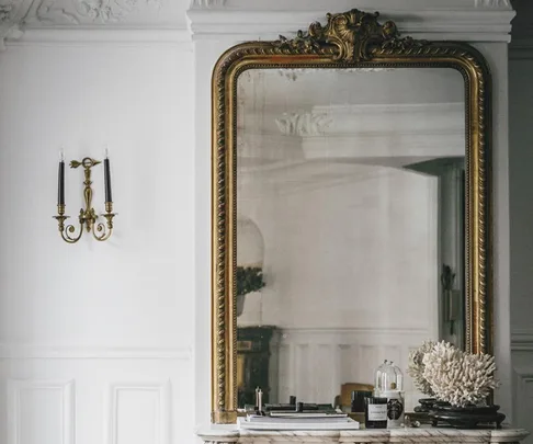 A large ornate gold-framed mirror above a marble mantel with decorative items and a brass wall sconce to the left.