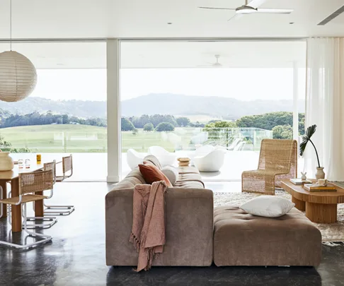 Modern living room with large windows, a brown couch, wicker furniture, and views of a green landscape and hills in the distance.