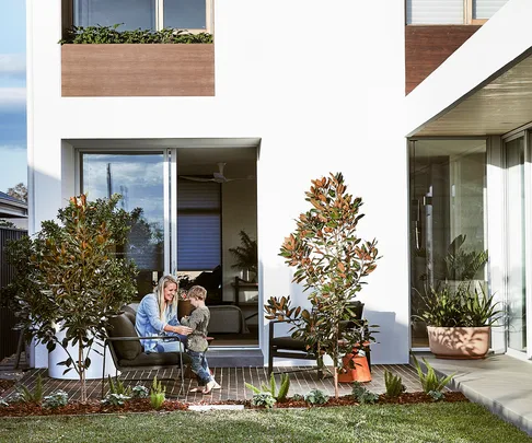 A woman and child sitting in a modern garden patio outside a white house with sliding glass doors and green plants.