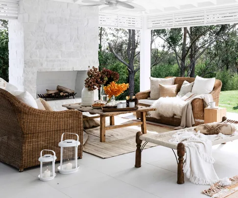 Outdoor patio with wicker furniture, a white stone fireplace, a wooden coffee table, and decorative lanterns, surrounded by greenery.