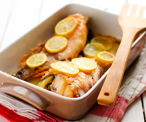 Baked fish topped with lemon slices in a beige ceramic dish next to a wooden spatula and red and white kitchen towels.