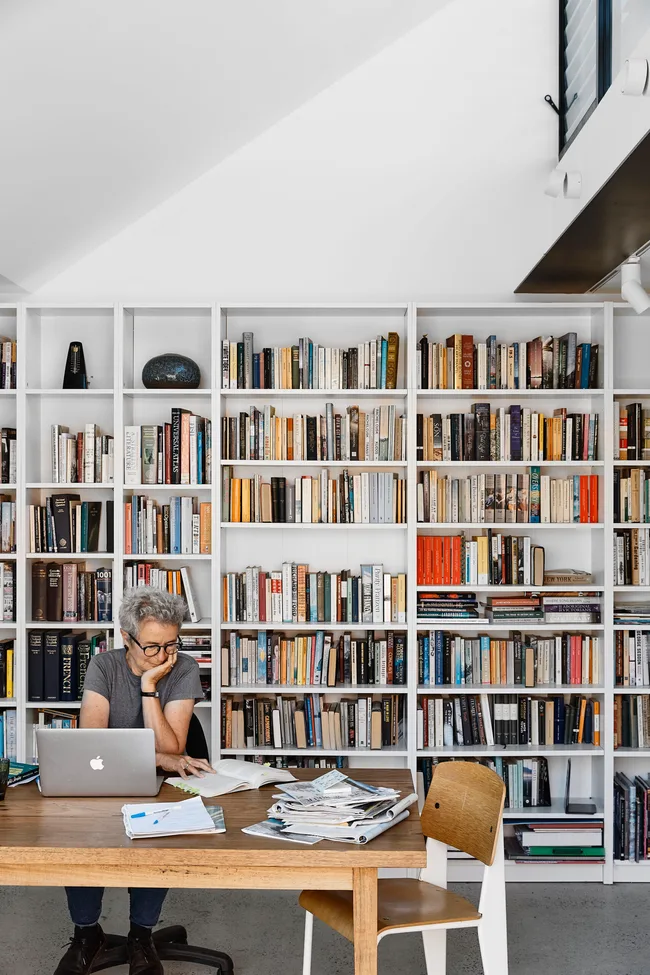 Person with gray hair working at a wooden desk, surrounded by books and papers, in a room with a large bookshelf.