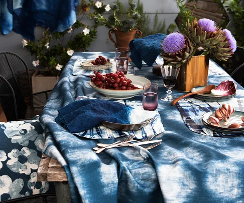 A beautifully set dining table with a blue and white tablecloth, purple flowers, grapes, garlic, and glasses of wine and water.