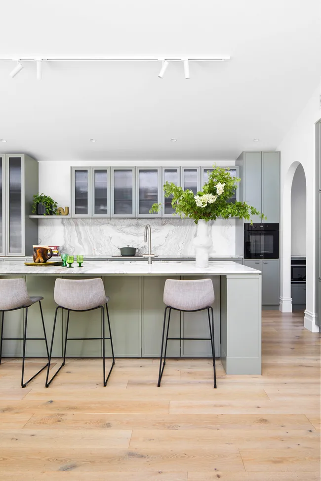 Modern kitchen with light gray cabinets, marble backsplash, island with stools, and a large vase with greenery.