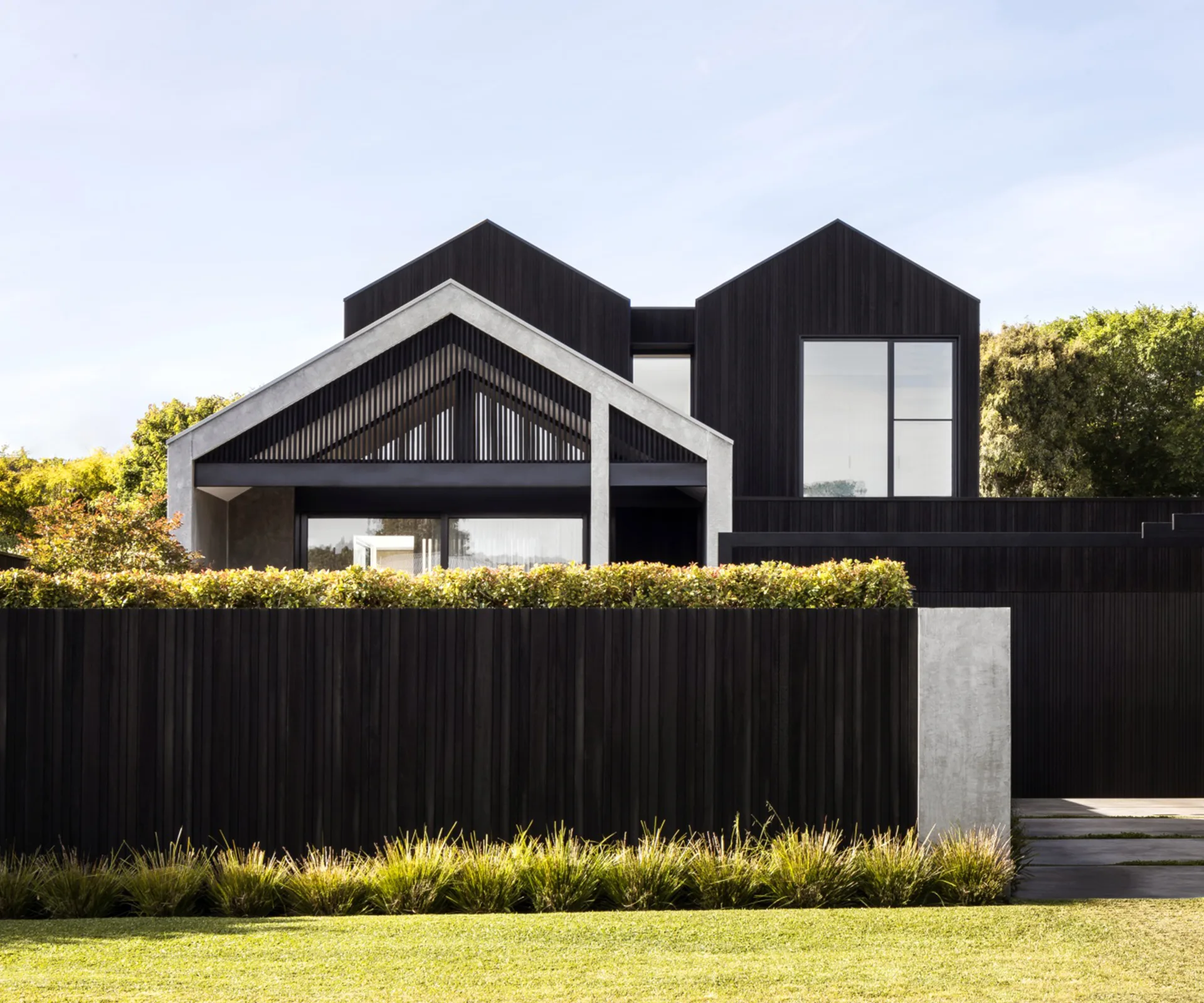 Modern house with dark exterior, large windows, and a tall fence, set behind a well-maintained lawn and green shrubs.