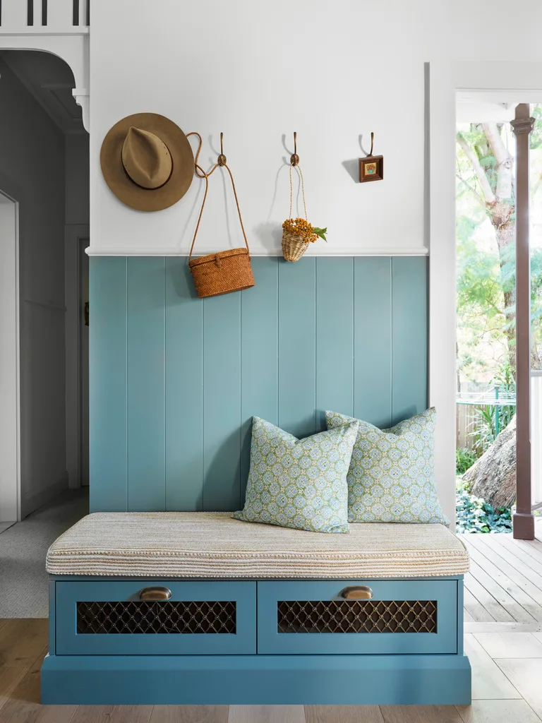 Cozy entryway with blue bench, pillows, hat, and baskets on hooks, adjacent to a window with a garden view.