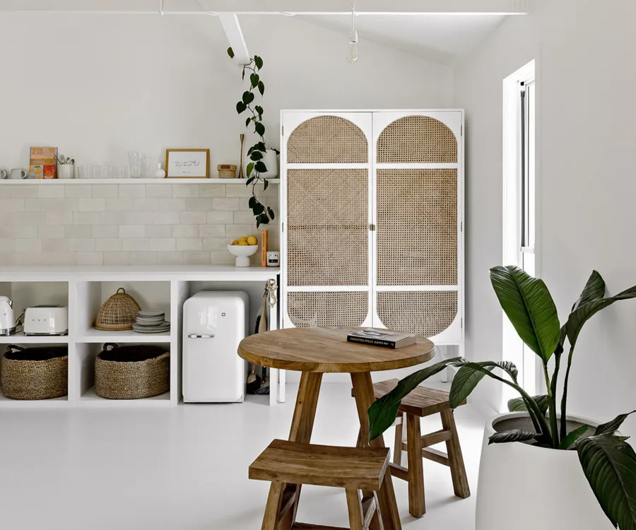 Alt text: "Minimalist kitchen with wooden table, stools, woven cabinet, and potted plant."