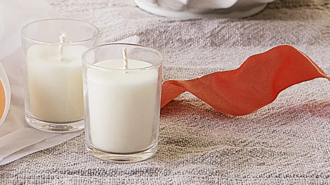Two white soy candles in glass containers on a textured surface with a red ribbon beside them.